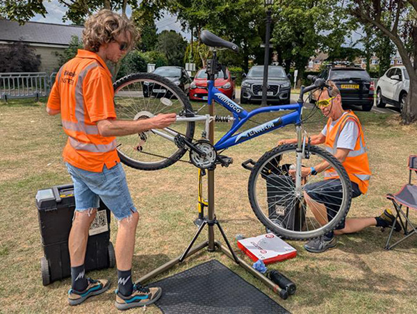 2 men from the Dr Bike team, dressed in orange hi-vis vests, fixing a bike
