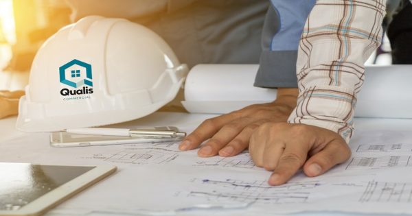 Hard hat with Qualis logo and two hands pointing at plans on a table